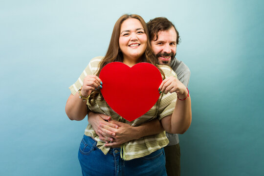 Hispanic couple smiling, standing against a blue background, woman holding a red heart, expressing love and togetherness