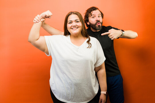 Empowered woman flexing arm with proud man, celebrating body positivity, strength, and confidence against orange background