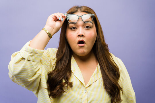 Plus size woman lifting eyeglasses on forehead, showing shock and astonishment on purple background