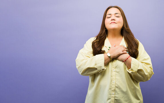 Plus size woman expressing gratitude, mindfulness, and peace, closing eyes, hands on chest feeling thankful and calm