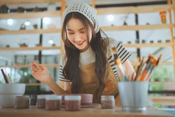 Happy Asian woman holding paintbrush and handcrafted ceramic bowl smiling at pottery workshop studio, cheerful artisan female artist showing handmade clay pottery product for small business art class © oatawa