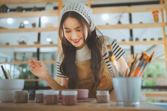 Happy Asian woman holding paintbrush and handcrafted ceramic bowl smiling at pottery workshop studio, cheerful artisan female artist showing handmade clay pottery product for small business art class