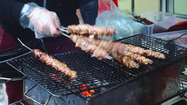 A person wearing gloves grills reddish-brown meat skewers over glowing embers at a busy Asian street food stall with supplies in the background.