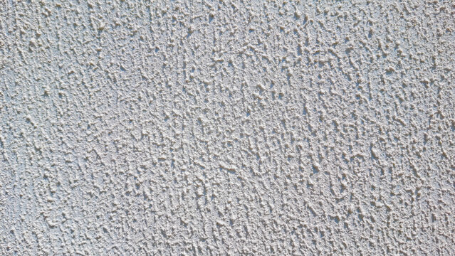 Coarse rough white stucco wall texture background with subtle sunlight casting soft shadows over the granular surface.