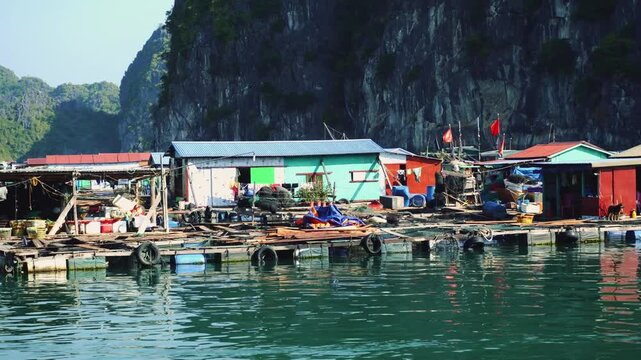 A colorful floating fishing village with wooden houses and boats on Ha Long Bay near Cat Ba Island, Vietnam, set against towering limestone karst mountains during daytime.