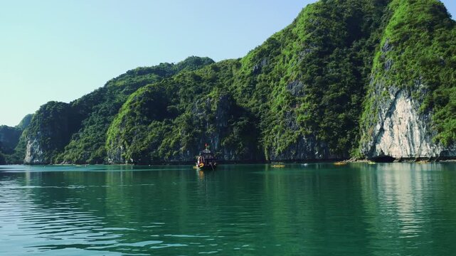 A dark multi-story tourist boat sails calmly among towering limestone karsts covered with lush green vegetation in the tropical seascape of Halong Bay, Vietnam, during daytime.
