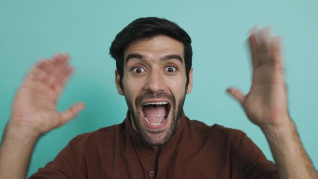 Excited young man with shocked expression opening his mouth wide and raising hands in celebration. Surprised hispanic guy celebrating victory or big news over plain blue background