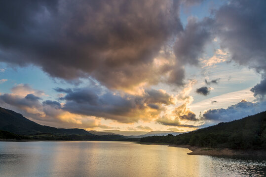 Scenic view of Ordunte reservoir at sunset surrounded by mountains under dark dramatic stormy clouds with warm light reflections on calm water