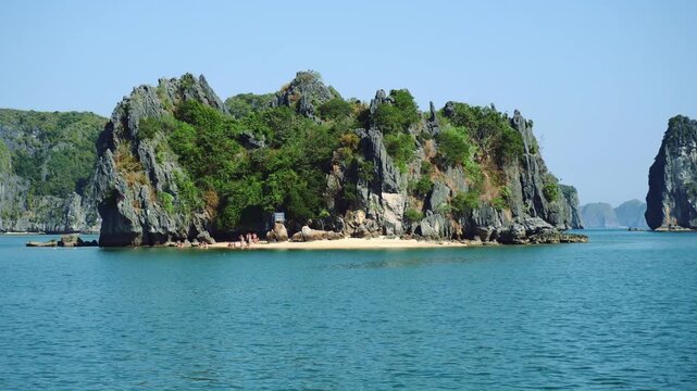 A scenic daytime view of a large rocky island with dense tropical vegetation and a sandy beach in Halong Bay, Vietnam, surrounded by calm turquoise waters and nearby islands.