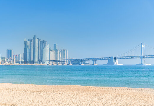 a scenic view of a modern city skyline with a large bridge spanning over the ocean, against a clear blue sky. The foreground features a sandy beach. in Busan, South Korea