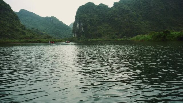 Tourists boating on a limestone karst lake surrounded by steep, jungle-covered mountains in the Ninh Binh region of Vietnam during daytime.