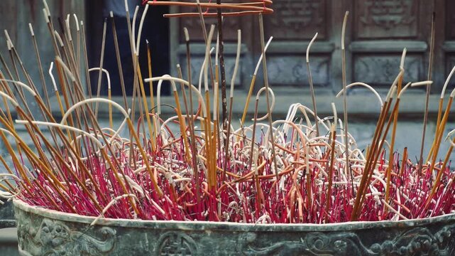 A stone planter filled with reddish foliage and burning incense sticks emits smoke in the courtyard of a traditional temple in urban Vietnam during daytime.