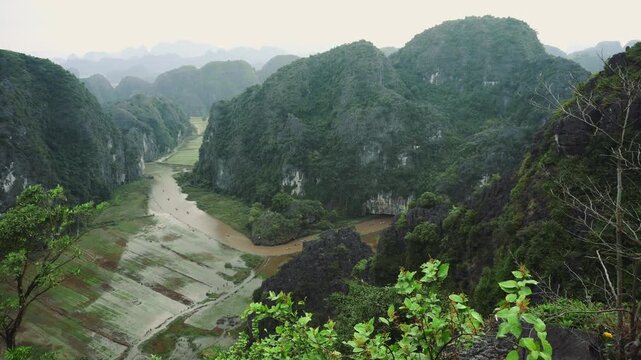 A panoramic daytime view of terraced rice fields and a winding river surrounded by limestone karst mountains in Hang Mua, Ninh Binh, Vietnam.
