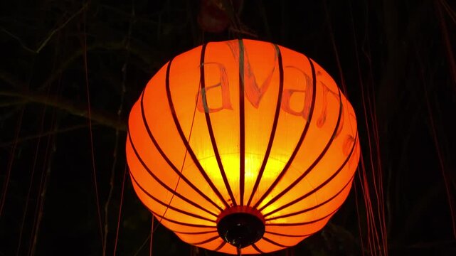 A large circular paper lantern glowing orange-yellow hangs outdoors at nighttime in the town of Hoi An, Vietnam, showcasing traditional craftsmanship and cultural heritage.