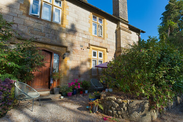 A charming, sunny old stone cottage with yellow walls, green foliage, and potted plants under a clear blue sky, suitable for architectural or travel themes - Kenegie Manor, Cornwall, UK © Robert