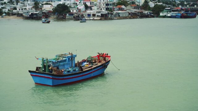 A large motorized fishing vessel with blue hull and red trim operates near a densely populated coastal village in Nha Trang, Vietnam during daytime.