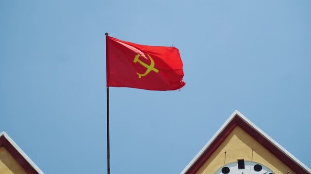 A red Soviet Union flag with a yellow hammer and sickle symbol waves on a flagpole against a clear blue sky in front of a building with peaked roof and arched windows.