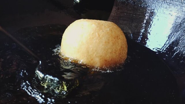 A large doughnut-like pastry is being fried in a pan with hot oil indoors in a Vietnamese kitchen during daytime. Close-up of cooking process without people.