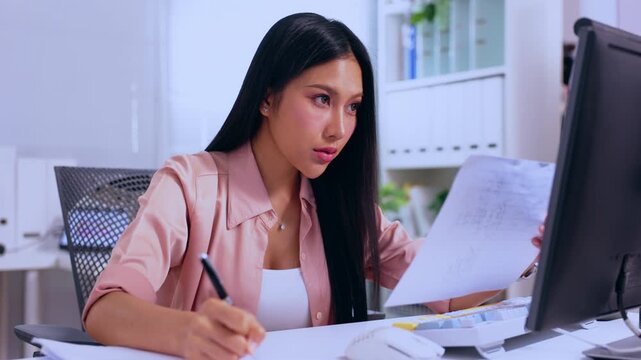Asian woman office paperwork computer desk pen business document work serious professional writing notes reviewing files modern workplace focused businesswoman concentration