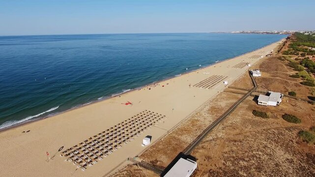 Drone footage panning slowly over the beautiful long sandy beach of vilamoura in the algarve, portugal, showing calm blue ocean waves, beach chairs, and sunbathers relaxing on a sunny day