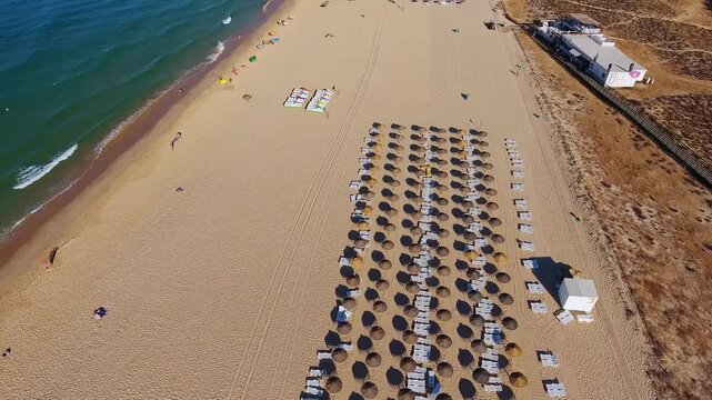 High aerial view flying along a beautiful sandy beach with rows of straw umbrellas and sunbeds next to the turquoise ocean waves, a perfect summer destination in vilamoura, algarve, portugal