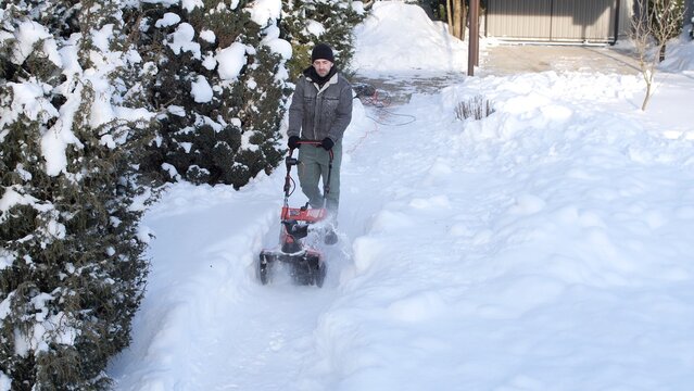 Man operating an electric snow blower, clearing deep snow from a residential pathway next to evergreen bushes during a cold winter day