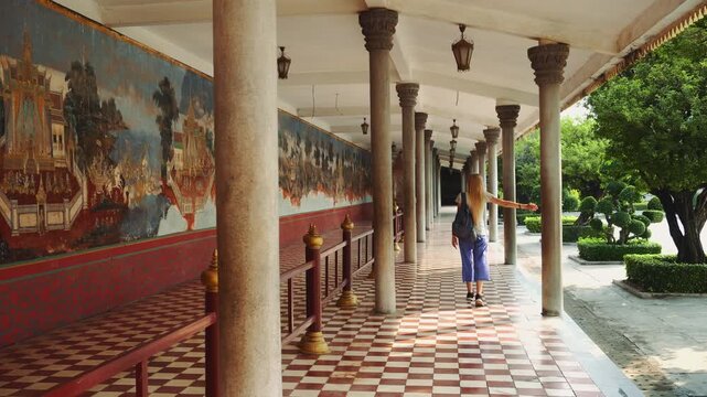 A blonde woman with a backpack walks along a long covered colonnade adorned with murals at the Royal Palace in Phnom Penh, Cambodia during daytime.