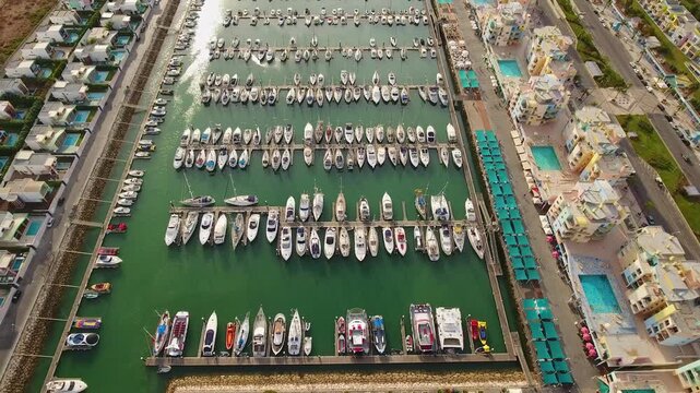 Aerial drone shot flying over the beautiful albufeira marina in portugal, revealing numerous luxury boats and yachts moored in the calm green water surrounded by colorful resort buildings