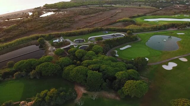 High angle drone footage capturing a modern water purification and wastewater treatment plant situated next to a beautiful green golf course and the coastline, showcasing sustainable infrastructure