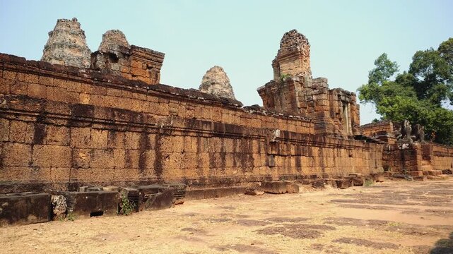Wide daytime view of ancient reddish-brown brick ruins with massive retaining walls and weathered towers at Angkor Wat archaeological site in Cambodia.