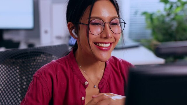 Asian woman with headset eyeglass in office at computer, remote worker calm communication, smiling online meeting in modern workspace with plant screen background