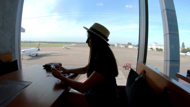 Woman in straw hat gazes out airport window, observing aircraft movement on the runway, capturing the anticipation of travel and the dynamic atmosphere of aviation in a modern terminal setting