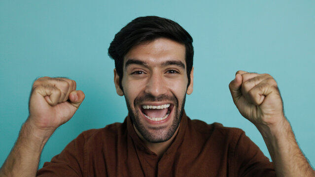 Excited young man with shocked expression opening his mouth wide and raising hands in celebration. Surprised hispanic guy celebrating victory or big news over plain blue background