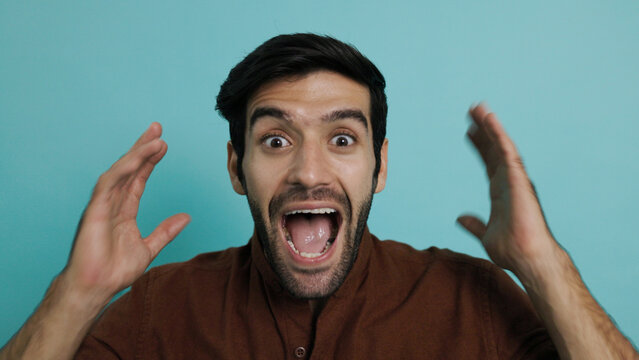 Excited young man with shocked expression opening his mouth wide and raising hands in celebration. Surprised hispanic guy celebrating victory or big news over plain blue background