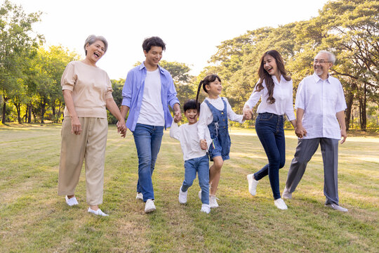 Happy multi-generational Asian family holding hands while walking through a sunny green park. Cheerful parents, children, and grandparents enjoying active leisure time together outdoors