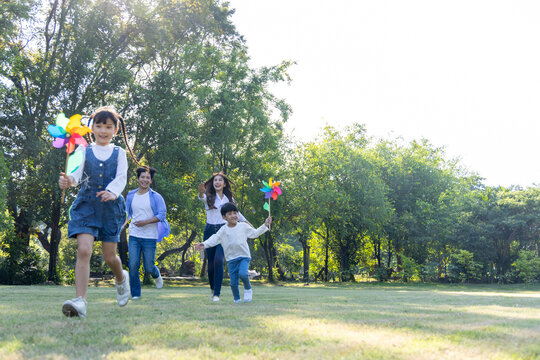 Cheerful Asian family playing together outdoors, with happy kids holding colorful pinwheels and running across a sunny green park while their smiling parents follow close behind