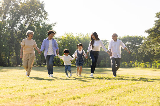 Joyful multi-generational Asian family holding hands while running together in a sunny park lawn. Happy parents, kids, and grandparents enjoying active quality leisure time outdoors