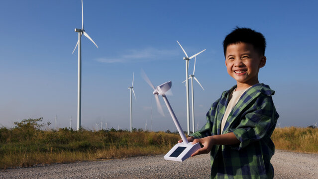 Happy young Asian boy holding a spinning toy wind turbine model in front of a real wind farm. Cheerful child learning about green renewable energy and eco-friendly power technology outdoors