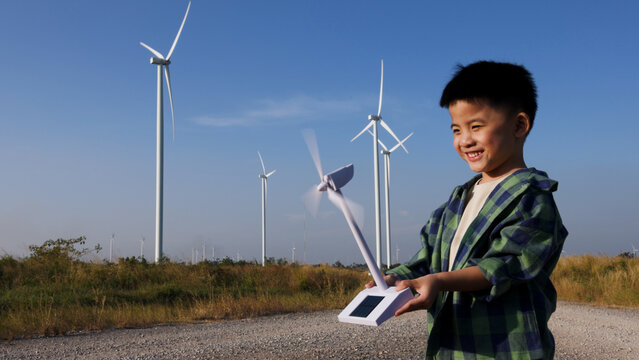 Happy young Asian boy holding a spinning toy wind turbine model in front of a real wind farm. Cheerful child learning about green renewable energy and eco-friendly power technology outdoors
