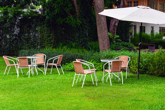 Garden patio set with white umbrella on lush green lawn. 
Outdoor seating area setup featuring woven wicker chairs, round tables, and large white patio parasol placed on vibrant green grass field.