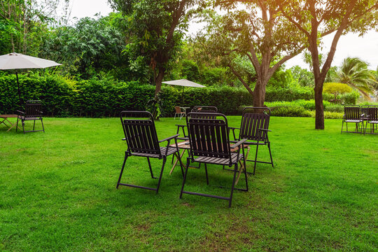 Outdoor patio seating sets on green lawn by river. Garden chairs and wooden tables on lush green grass field under shady trees in lakeside backyard, featuring peaceful riverfront landscape view.