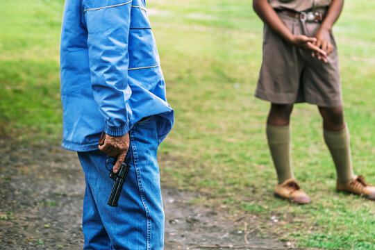 Sports referee holding starting pistol down by his side. Male sports official in jacket holding black starting pistol revolver downwards, standing by green grass field during a school athletic event.