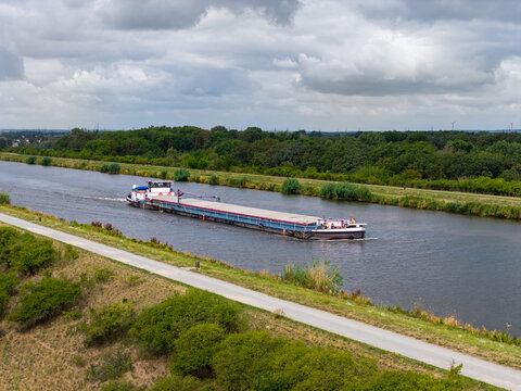 Cargo barge travels along Mittellandkanal through green landscape beneath dramatic summer clouds. Inland waterway supports freight transport, navigation and logistics across northern Germany