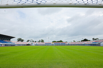 From inside the goal, this perspective shows white crossbar and net above, looking out over an empty outdoor football stadium featuring vibrant green grass and colorful grandstands under a cloudy sky. © Wonchalerm