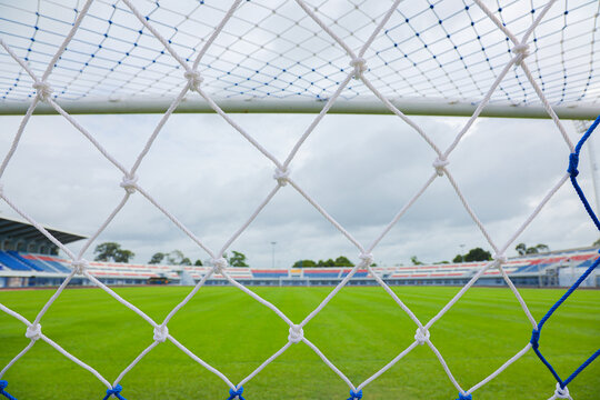 Selective focus of white soccer goal net in the foreground. Through the woven mesh, a blurred empty outdoor football stadium for vibrant green grass pitch under a naturally cloudy daytime sky scene.