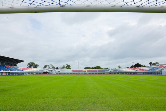 From inside the goal, this perspective shows white crossbar and net above, looking out over an empty outdoor football stadium featuring vibrant green grass and colorful grandstands under a cloudy sky.