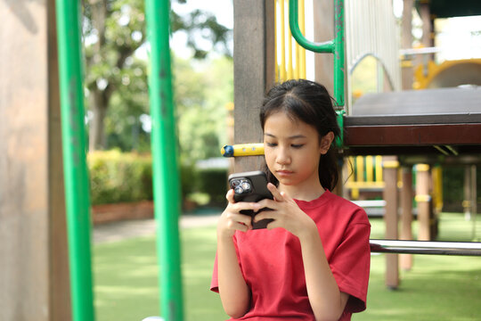 Selective focus outdoor shot captures Asian kid in a red shirt standing at a playground. The young girl is deeply engaged with a smartphone, highlighting modern childhood technology and screen time.
