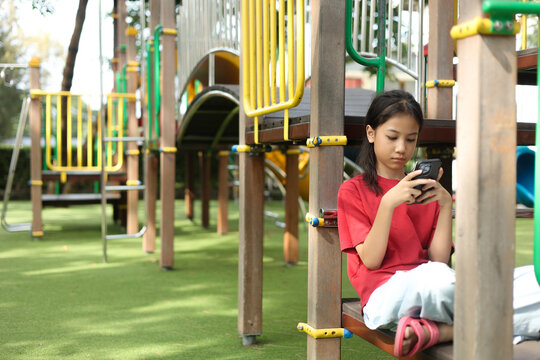 Outdoor shot with copy space captures Asian kid in a red shirt standing at a playground. The young girl is deeply engaged with a smartphone, highlighting modern childhood technology and screen time.