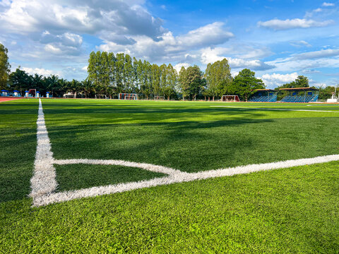 Wide angle view of green artificial grass soccer field with white corner line under a bright blue sky. Authentic outdoor stadium with stands and trees for sports competition and training backgrounds.