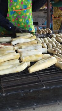 Vertical video with a right-to-left panning camera movement, showcasing a local vendor manually turning and rotating traditional Thai grilled cassava roots on a hot charcoal grill.